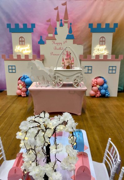 Princess-themed party setup with our beautifully decorated children's tables &  Chiavari chairs. The tables have our personalised boxes placed alongside our centrepieces. A pink castle-themed backdrop completes the magical atmosphere.